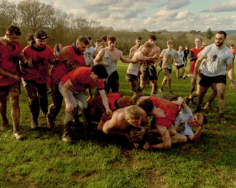 A large group of people wrestling on muddy grass, some wearing red t-shirts, trees and overcast sky in the background. 