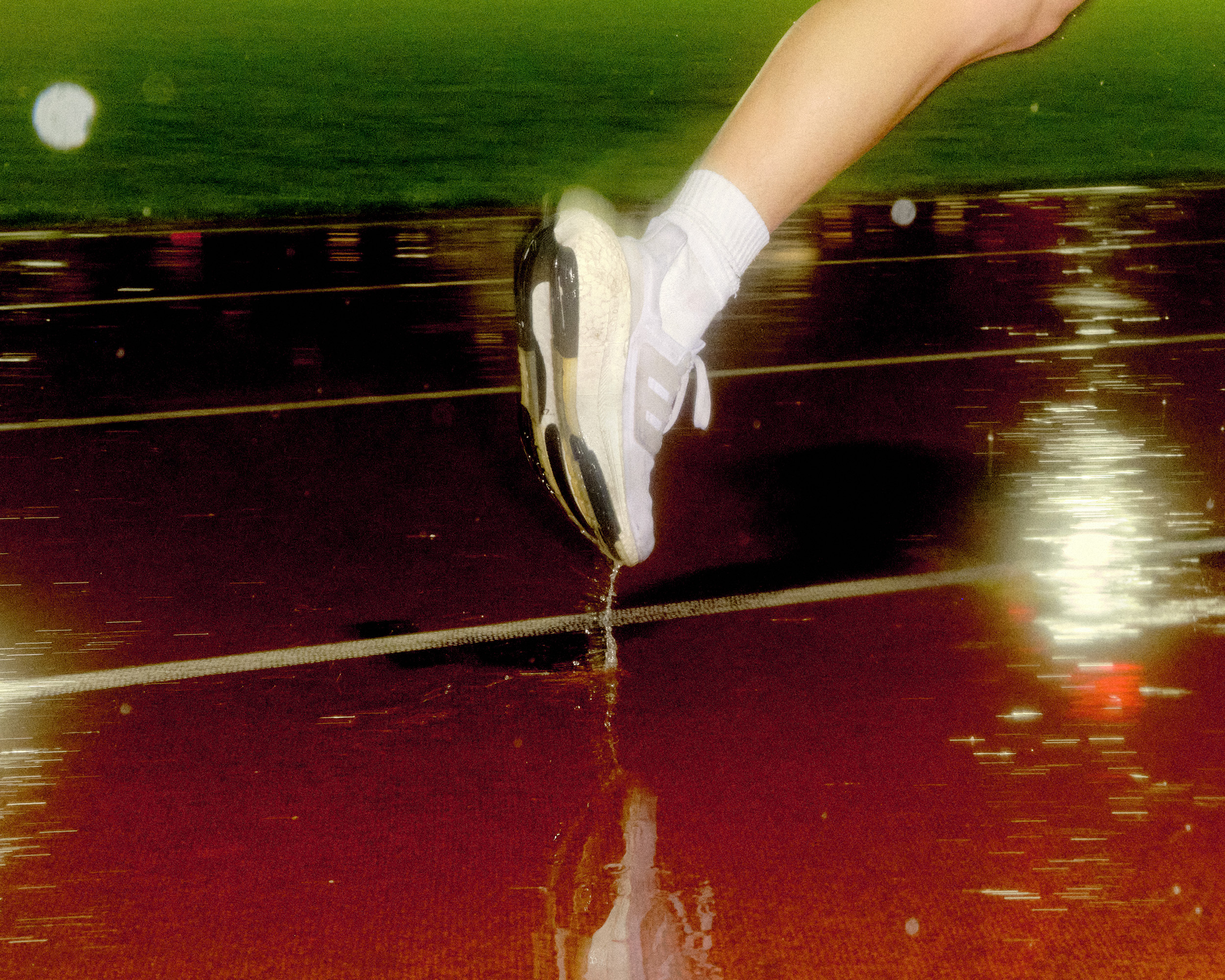 Runner's shoe stepping into a puddle on a wet track, creating a splash with reflected light and green background in motion blur.