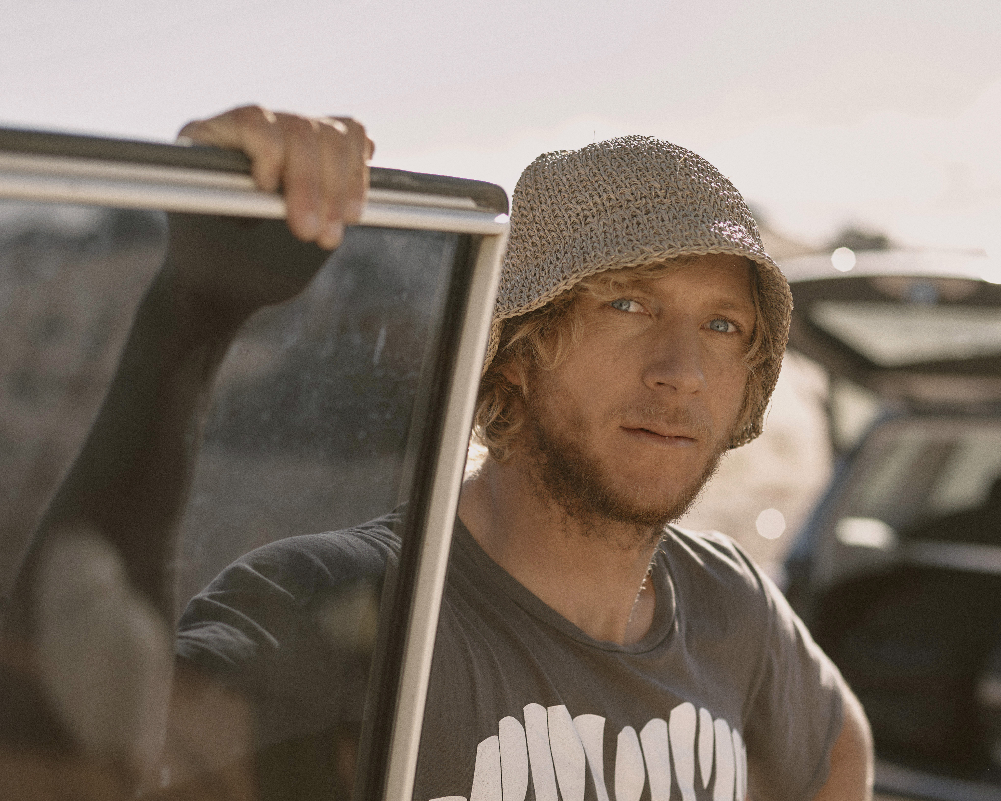 Man with a bucket hat stands next to an open car door. He is wearing a t-shirt with graphic text and has blond hair and a beard.