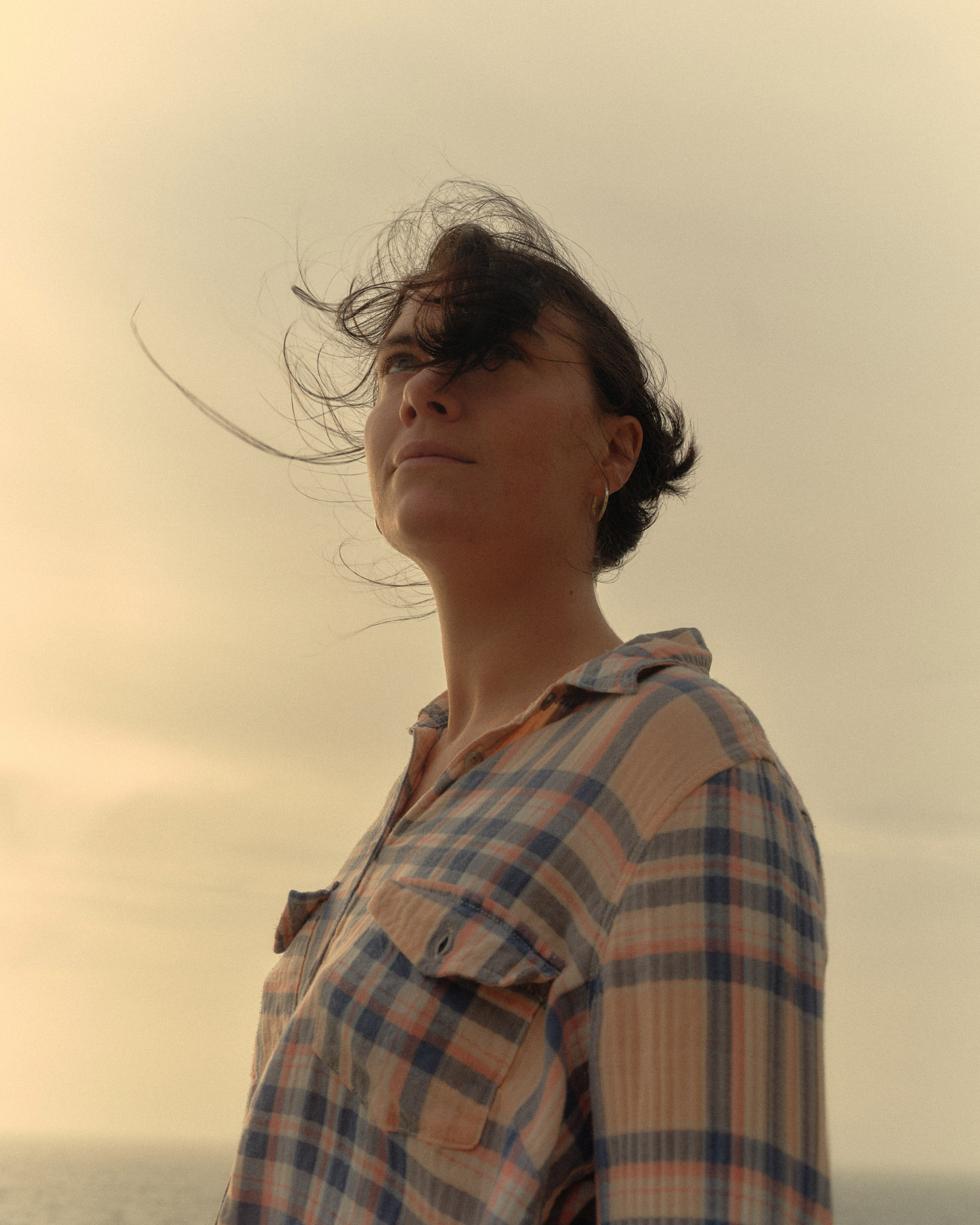 Woman in flannel shirt stands outdoors on a windy day with hair blowing across her face in a low-angle, eye-level portrait.