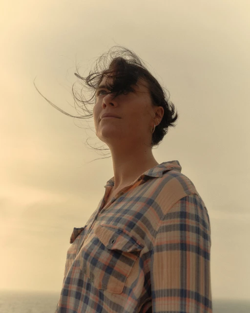 Woman in flannel shirt stands outdoors on a windy day with hair blowing across her face in a low-angle, eye-level portrait.
