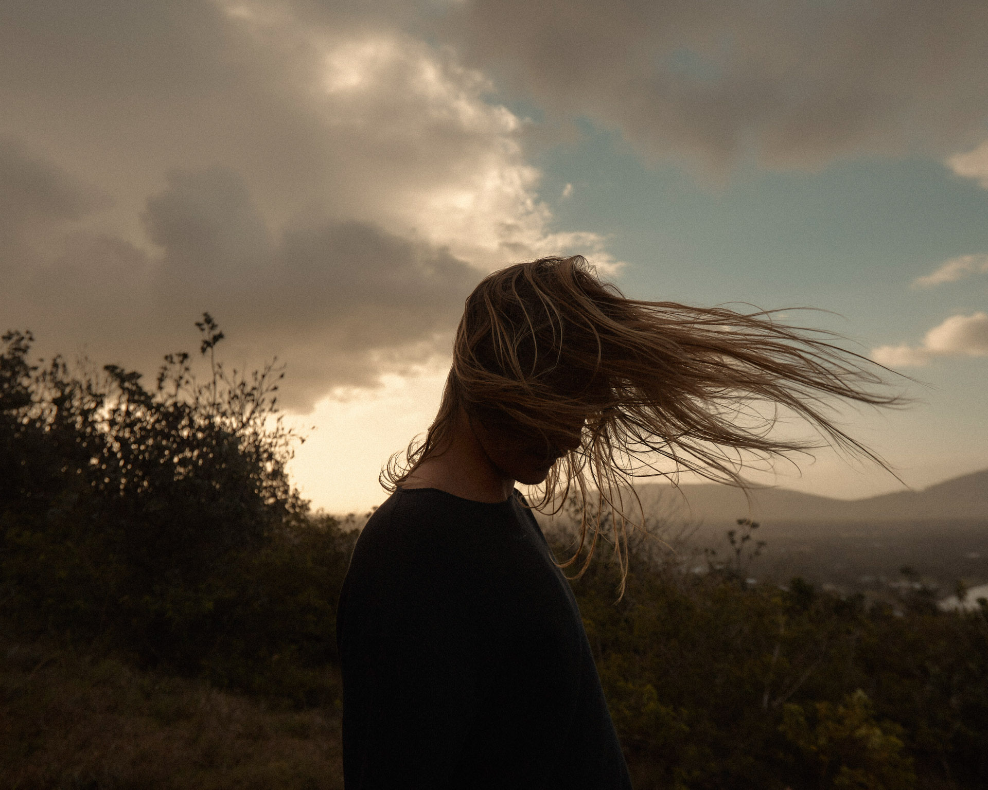 Woman with flowing hair silhouetted against a dramatic sky at sunset. Bush and hills in the background. Moody lighting, travel and nature theme.