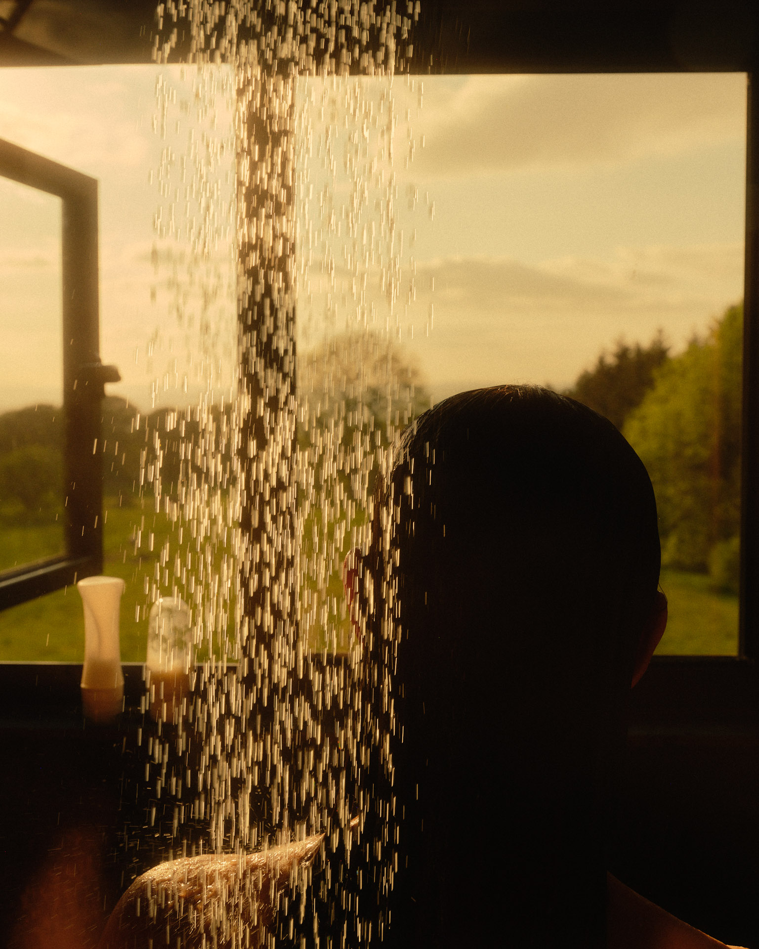 Person takes a shower in front of open window, overlooking a green rural landscape with trees under a light-yellow, clouded sky at daytime.