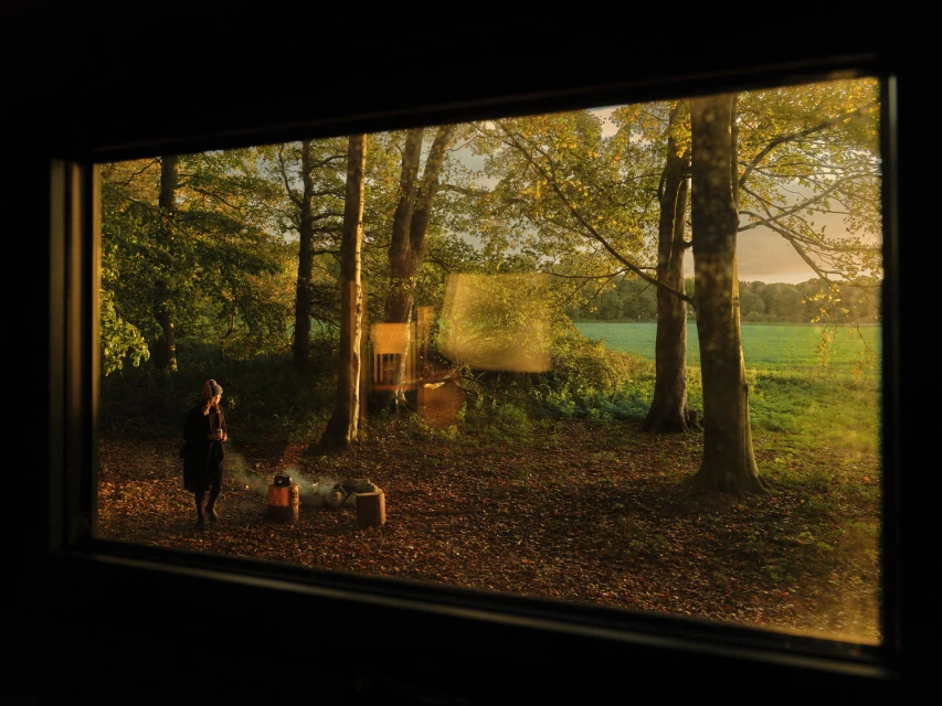 Woman in dark clothing stands near a small campfire in a leafy forest. A view framed through a dark window into a fall landscape.