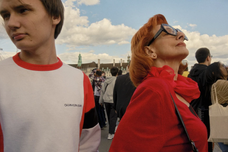 Outdoor image captures a young adult male and older adult female with bright red hair, against a backdrop of an urban scene and cloudy sky.