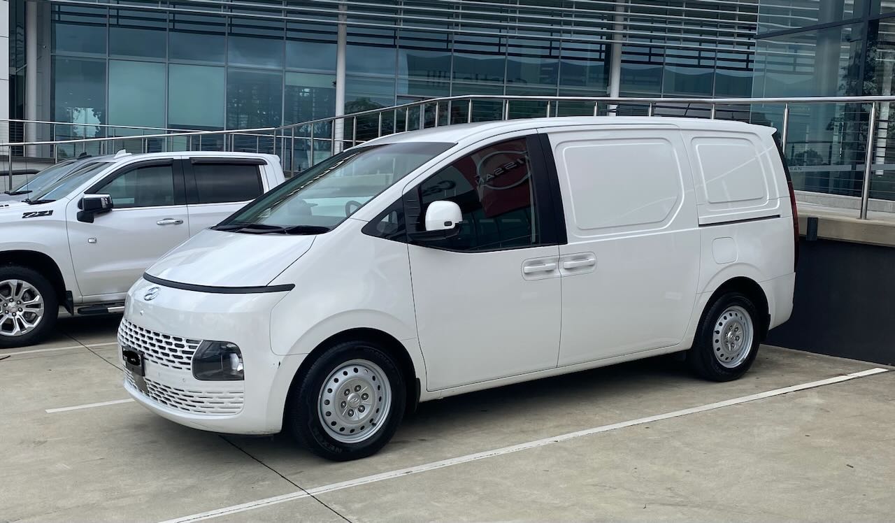 White Hyundai Staria Load parked outside a modern building on a sunny day, showcasing its sleek design and functionality.