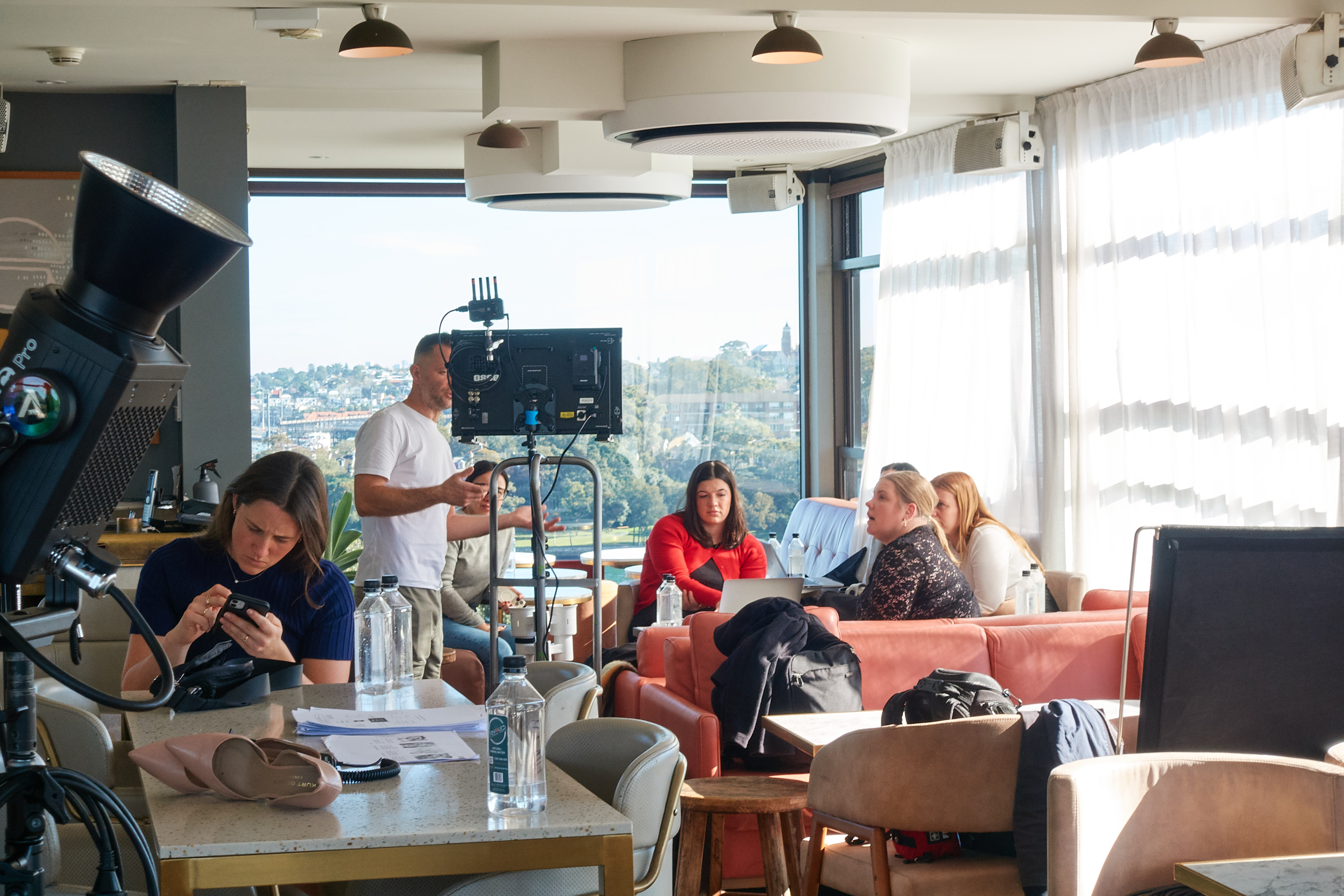 A group of people are seated indoors at tables, with film crew visible in the background. Large windows offer views of the urban landscape.