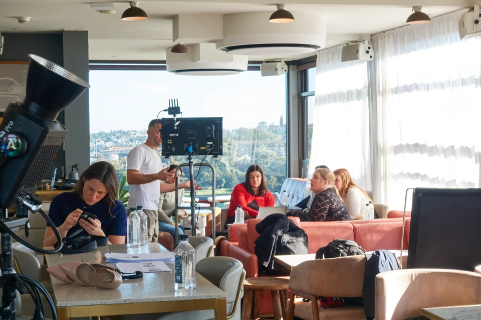 A group of people are seated indoors at tables, with film crew visible in the background. Large windows offer views of the urban landscape.