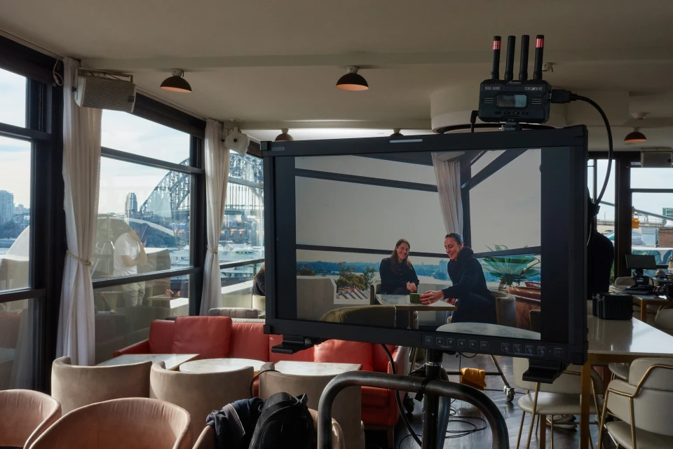 Two women are visible on a display screen indoors, with a partial view of a city visible in the background through large windows.