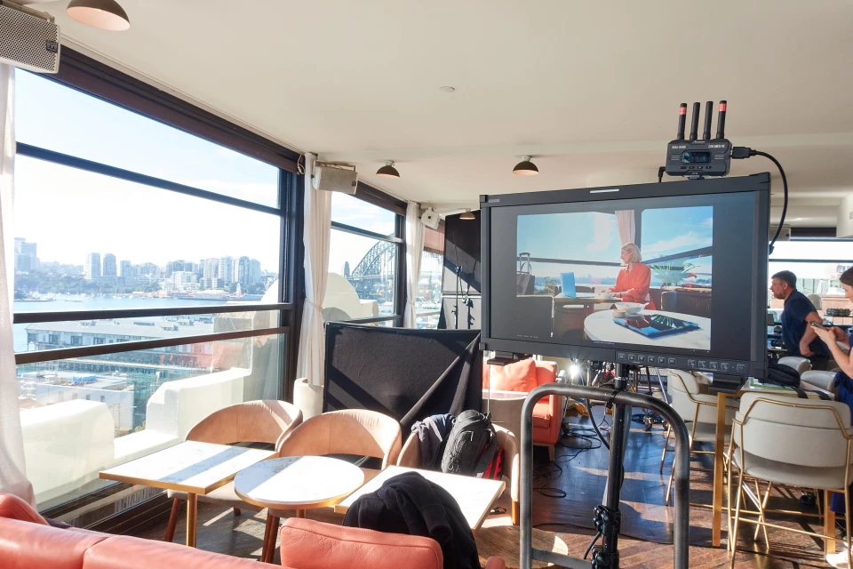 Indoor shot of a TV on a stand showing a woman on screen in a sunny room with an urban skyline view, along with chairs.
