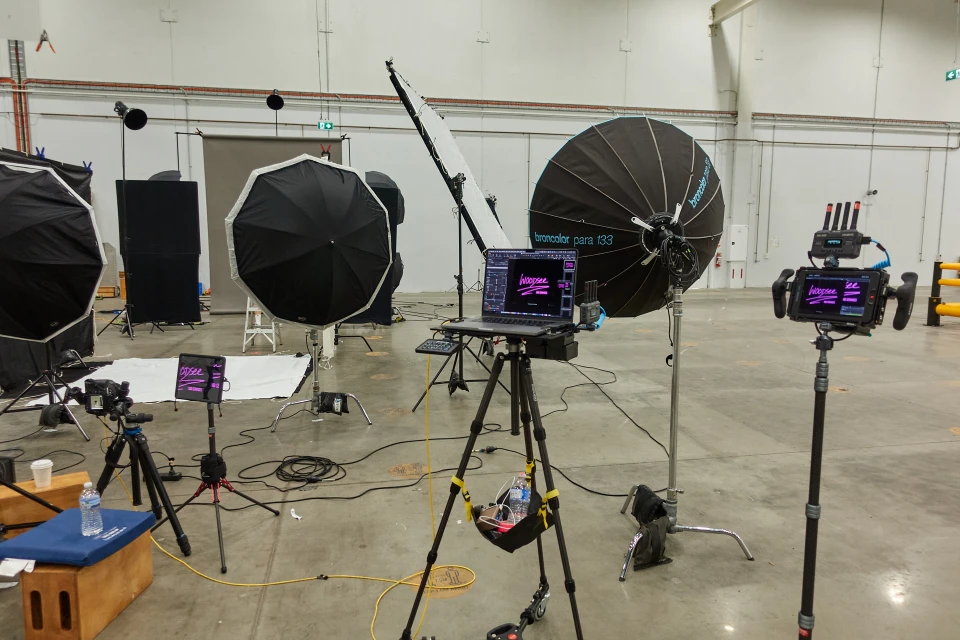 Photo studio setup with umbrellas, screens, cameras, and laptops on tripods, creating a backstage view of a professional photoshoot.