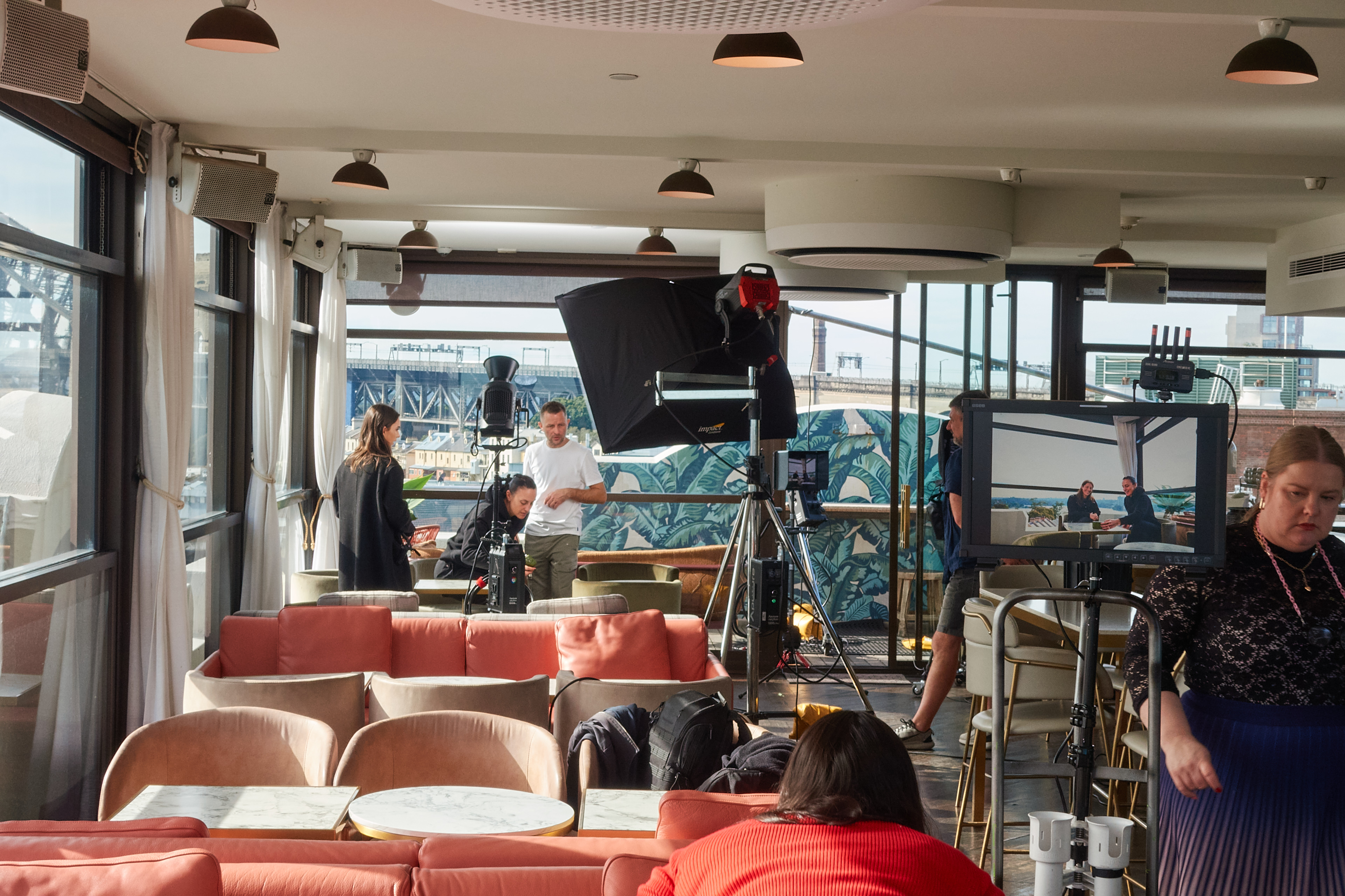 Photo shoot in an indoor space with a film crew, lighting equipment, coral-colored seating, and a cityscape visible in the background.