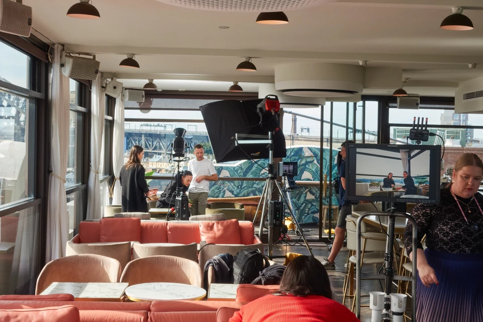 Photo shoot in an indoor space with a film crew, lighting equipment, coral-colored seating, and a cityscape visible in the background.