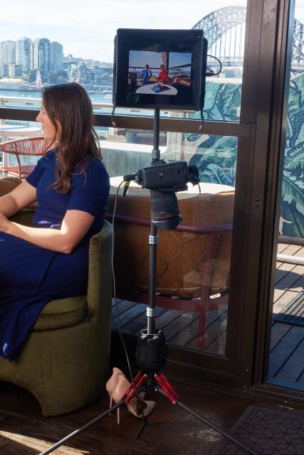 Woman sits near camera and monitor with city view. Camera on tripod, monitor displays a scene, and woman in blue dress is seated.