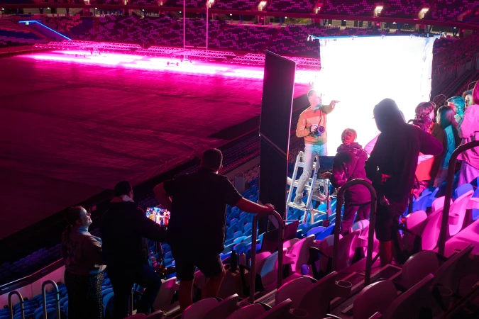 Filming on a stadium floor with a large group, camera crew, pink lighting, and equipment set up for a production with various people and lighting.