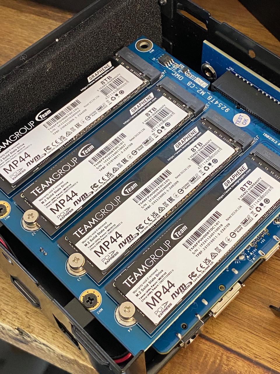 Internal view of a computer case with four TEAMGROUP MP44 NVMe M.2 solid state drives installed on a blue motherboard; close-up.