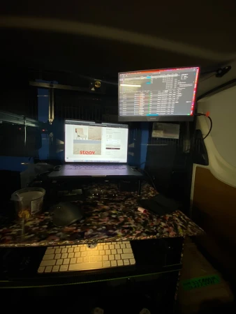 Mobile editing workstation setup inside a vehicle with dual monitors, laptop, and keyboard on a patterned desk.