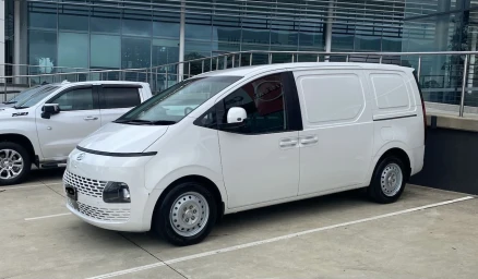White cargo van parked in an asphalt lot outside a modern commercial building with glass exterior walls and metal railings.