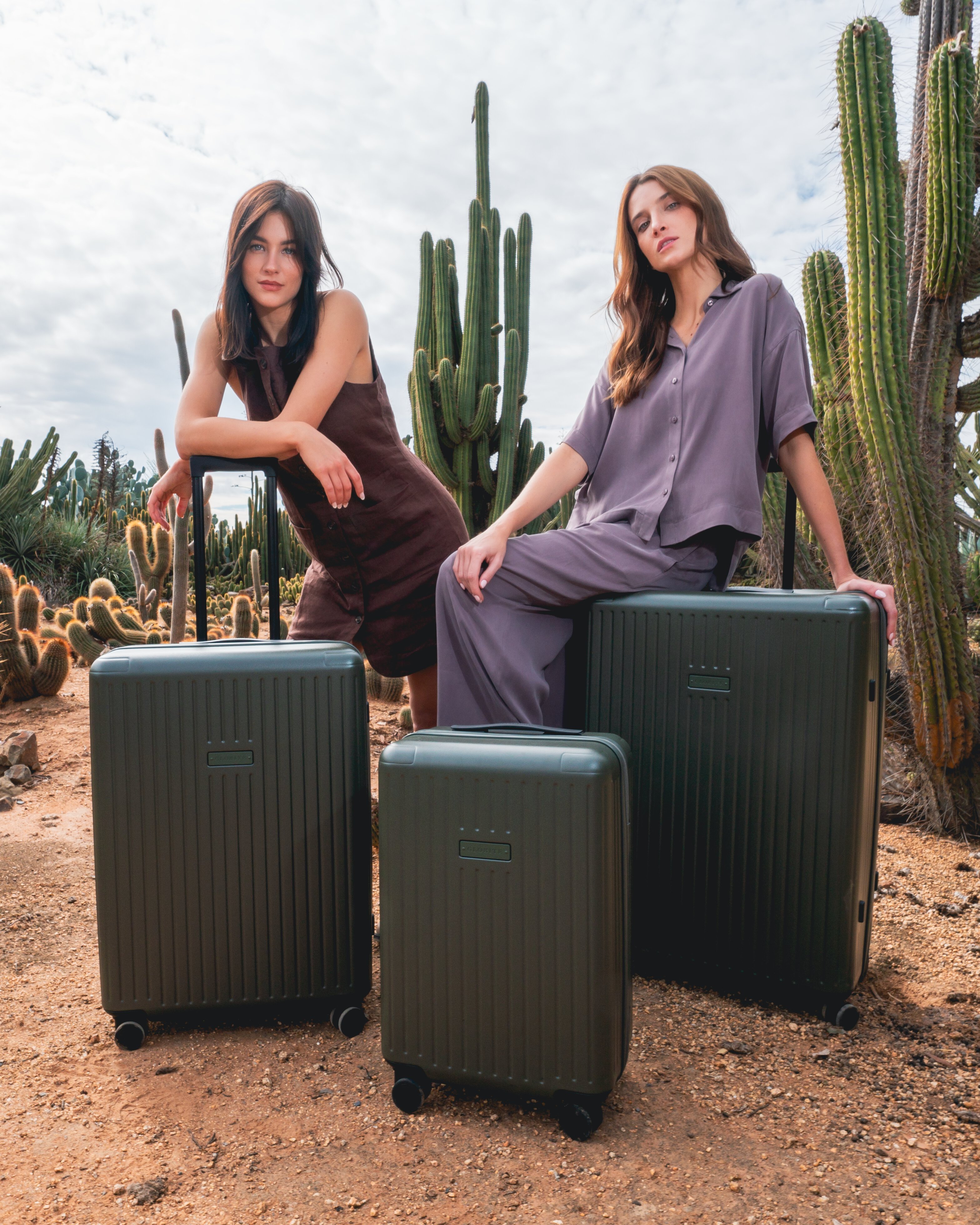Two women pose with olive green suitcases in an arid landscape with cacti under a sunny sky, showcasing travel accessories.
