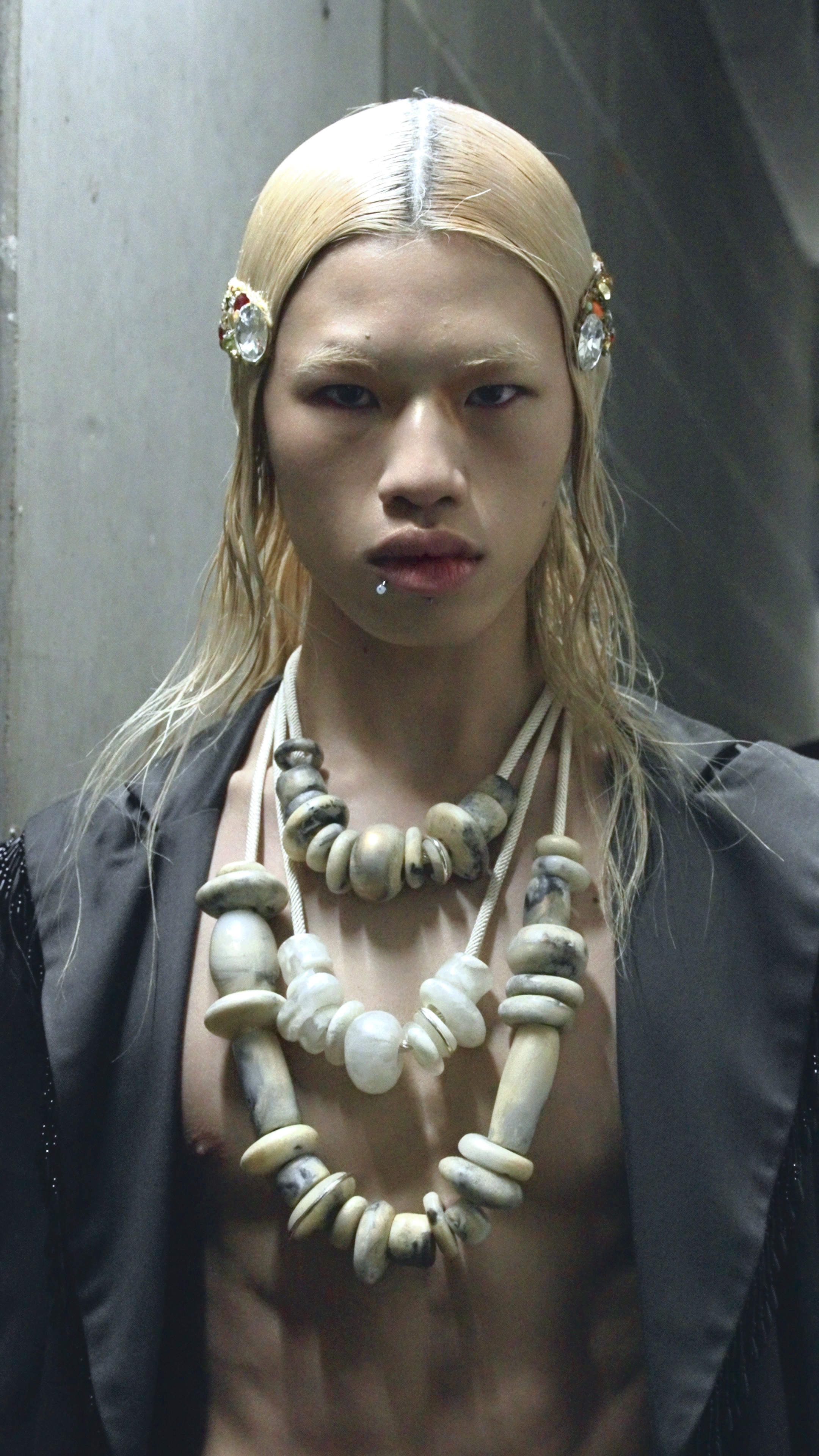 Striking close-up of a bare-chested fashion model with blond hair and multiple layered necklaces, set against a neutral grey background.