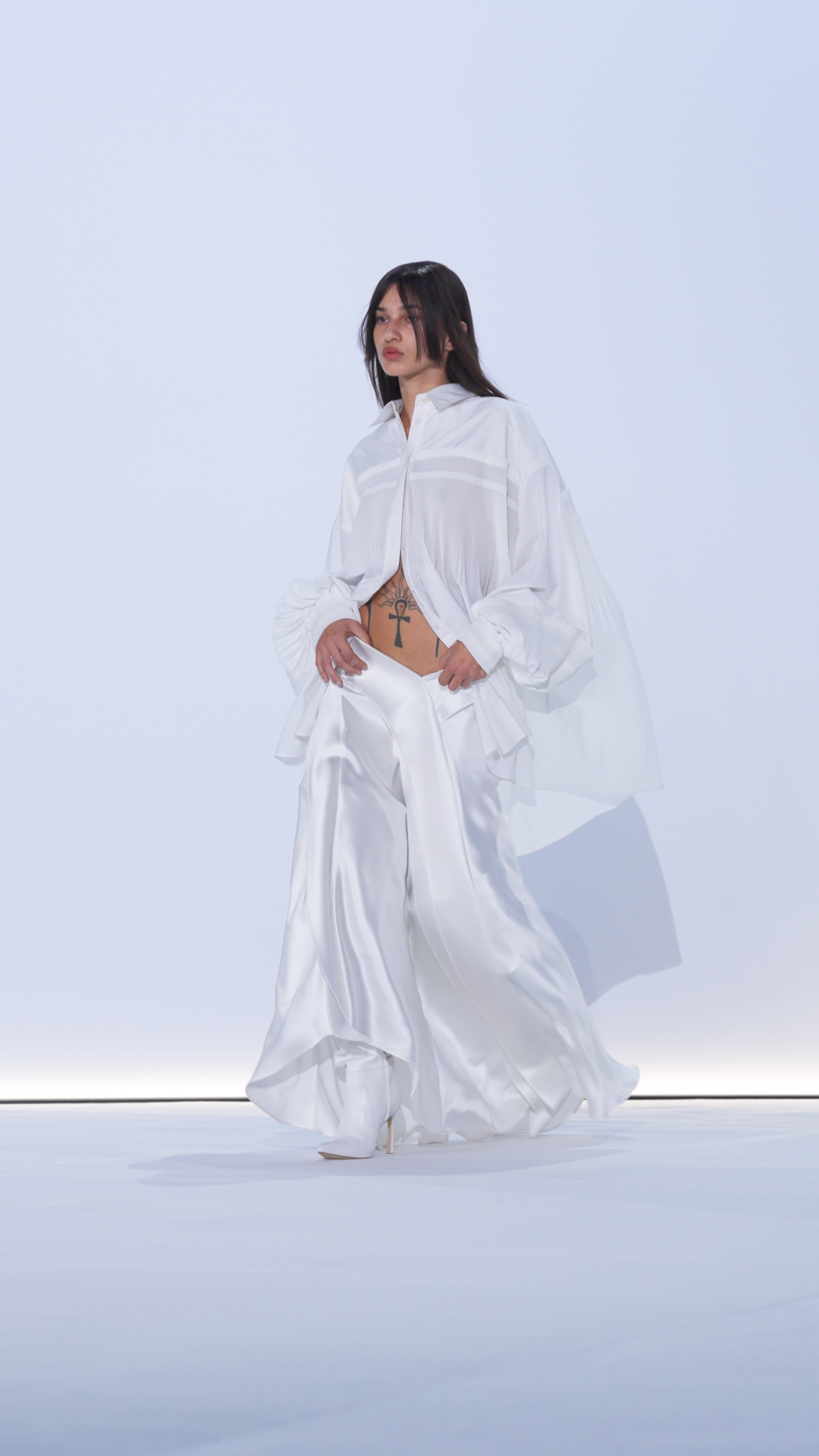 Runway model in white, flowing outfit walks on a white platform in a brightly lit fashion show, showcasing clothing design.