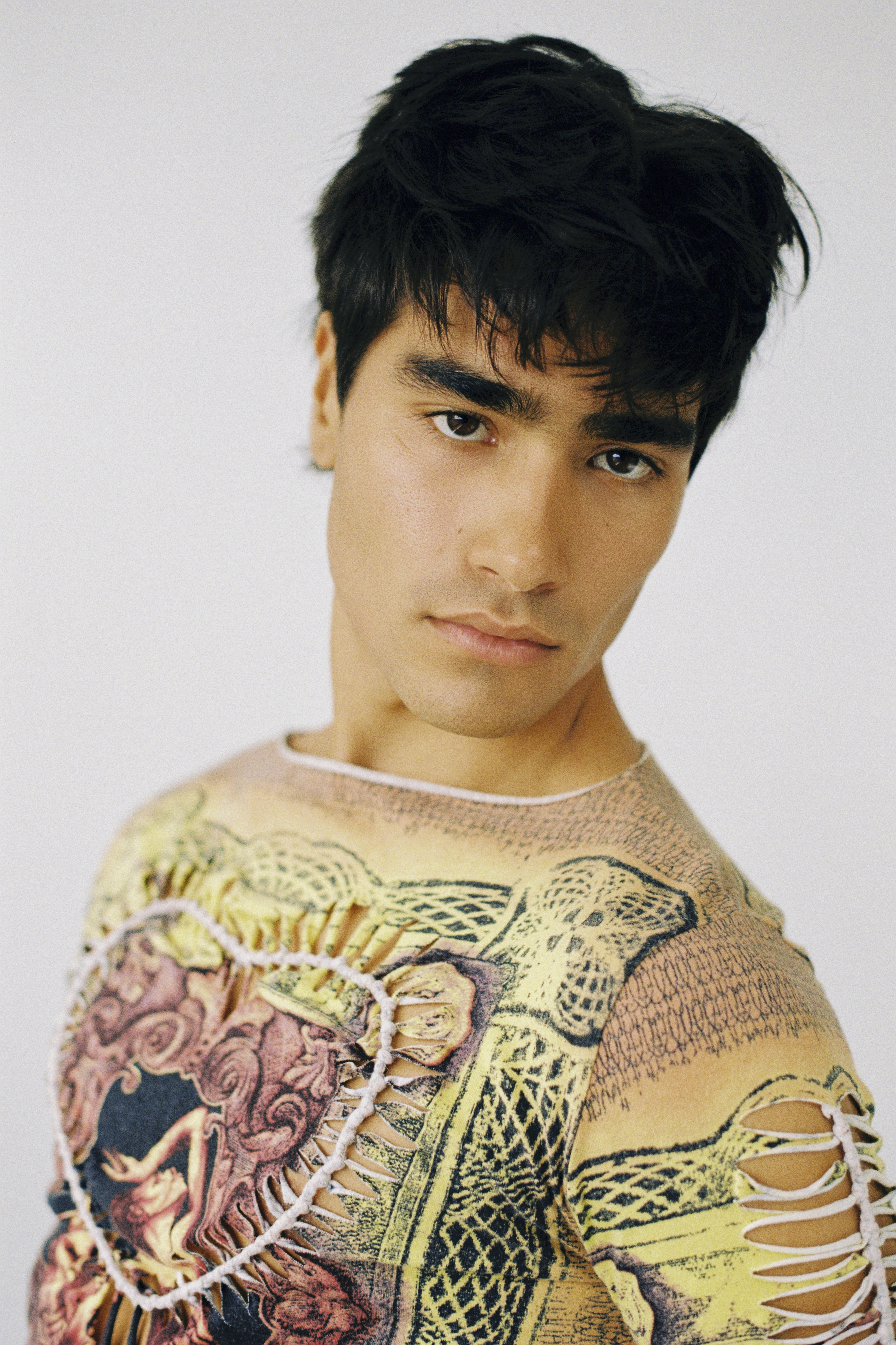 Portrait of an adult male with dark hair, wearing a graphic print shirt with distressed details against a solid white background.