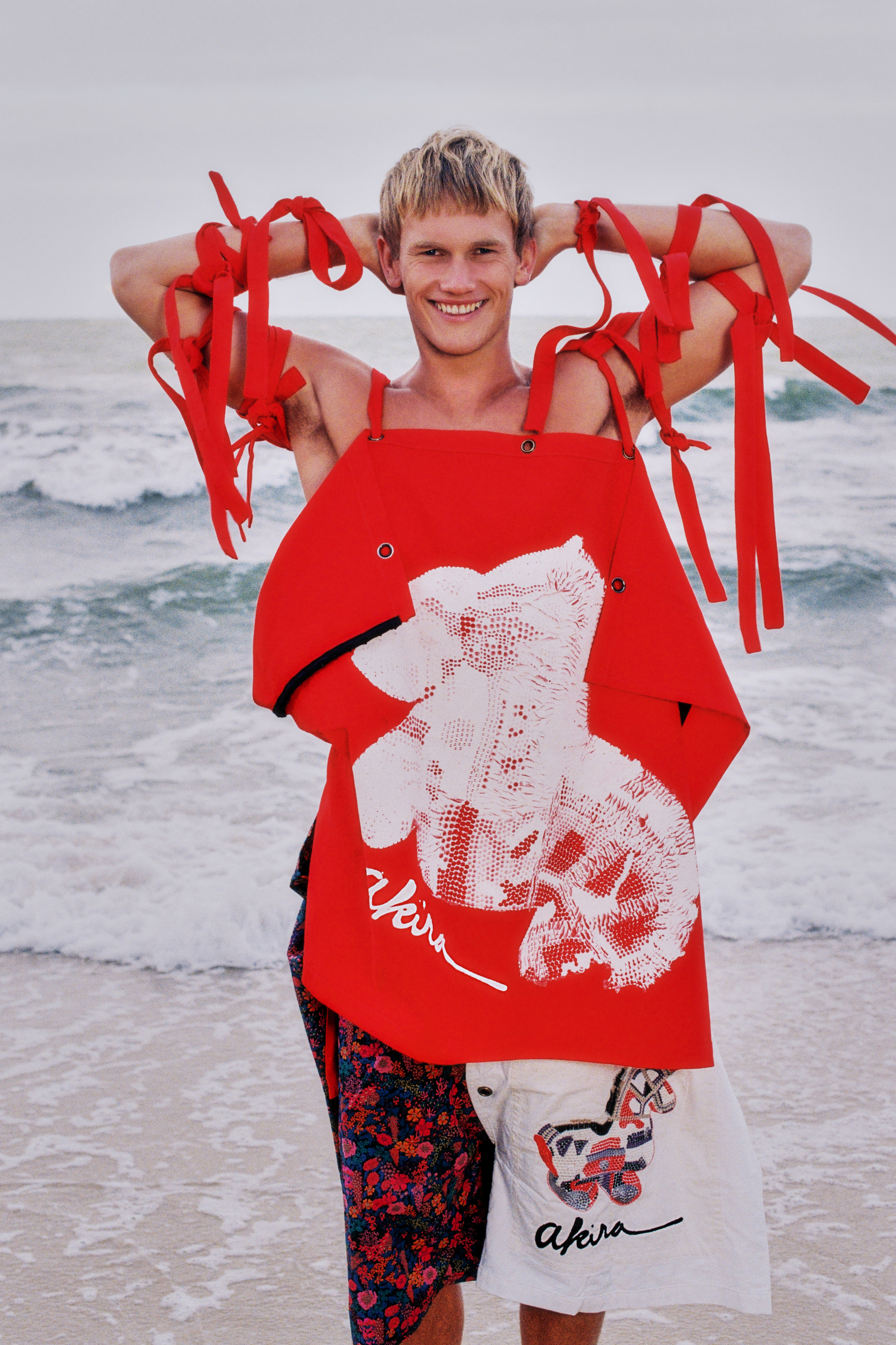 Smiling man poses on beach in avant-garde fashion, wearing deconstructed clothing with red ribbons and printed floral and graphic shorts.