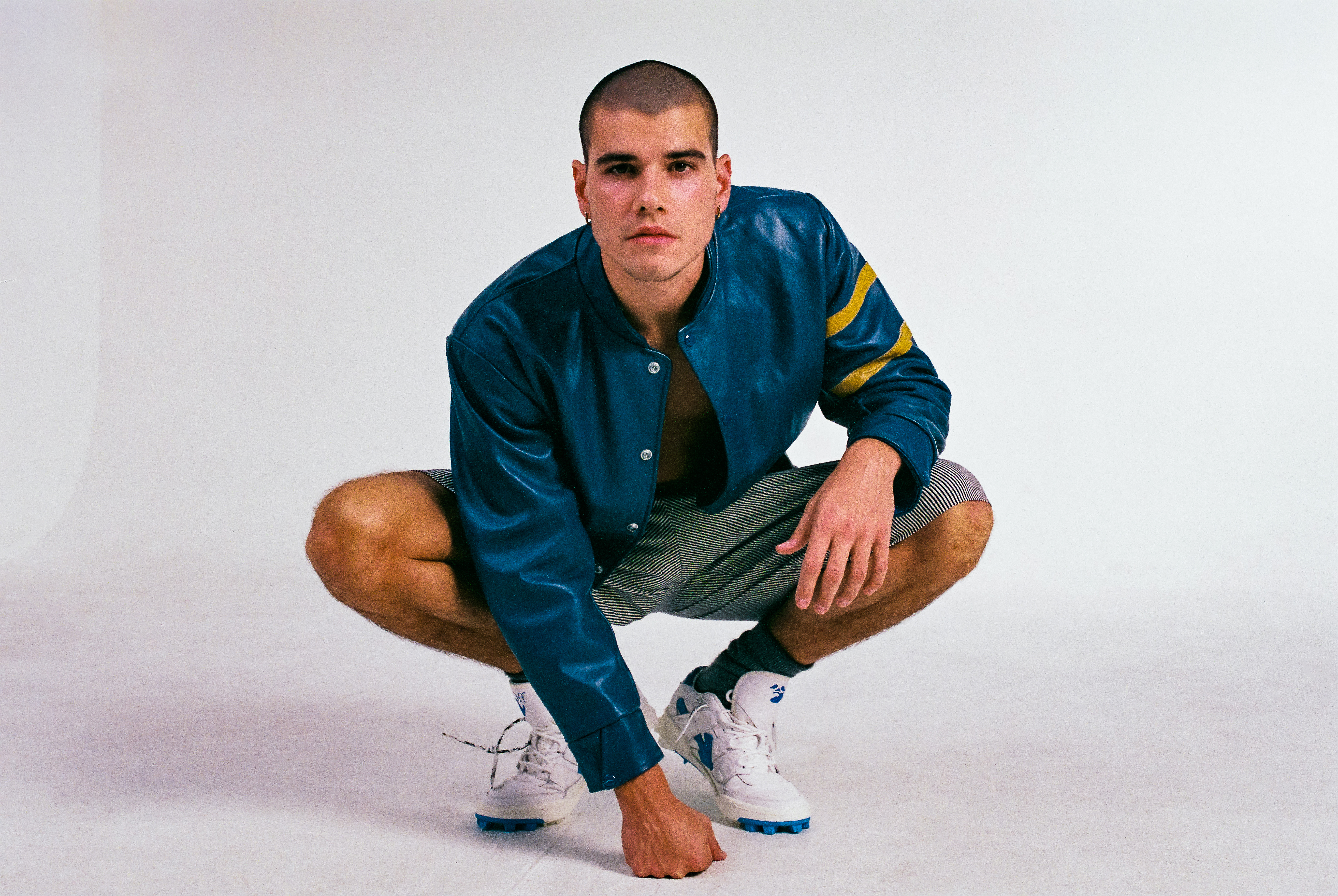 Male model in a blue jacket poses in a crouching position against a white backdrop, showcasing fashion apparel and athletic footwear.