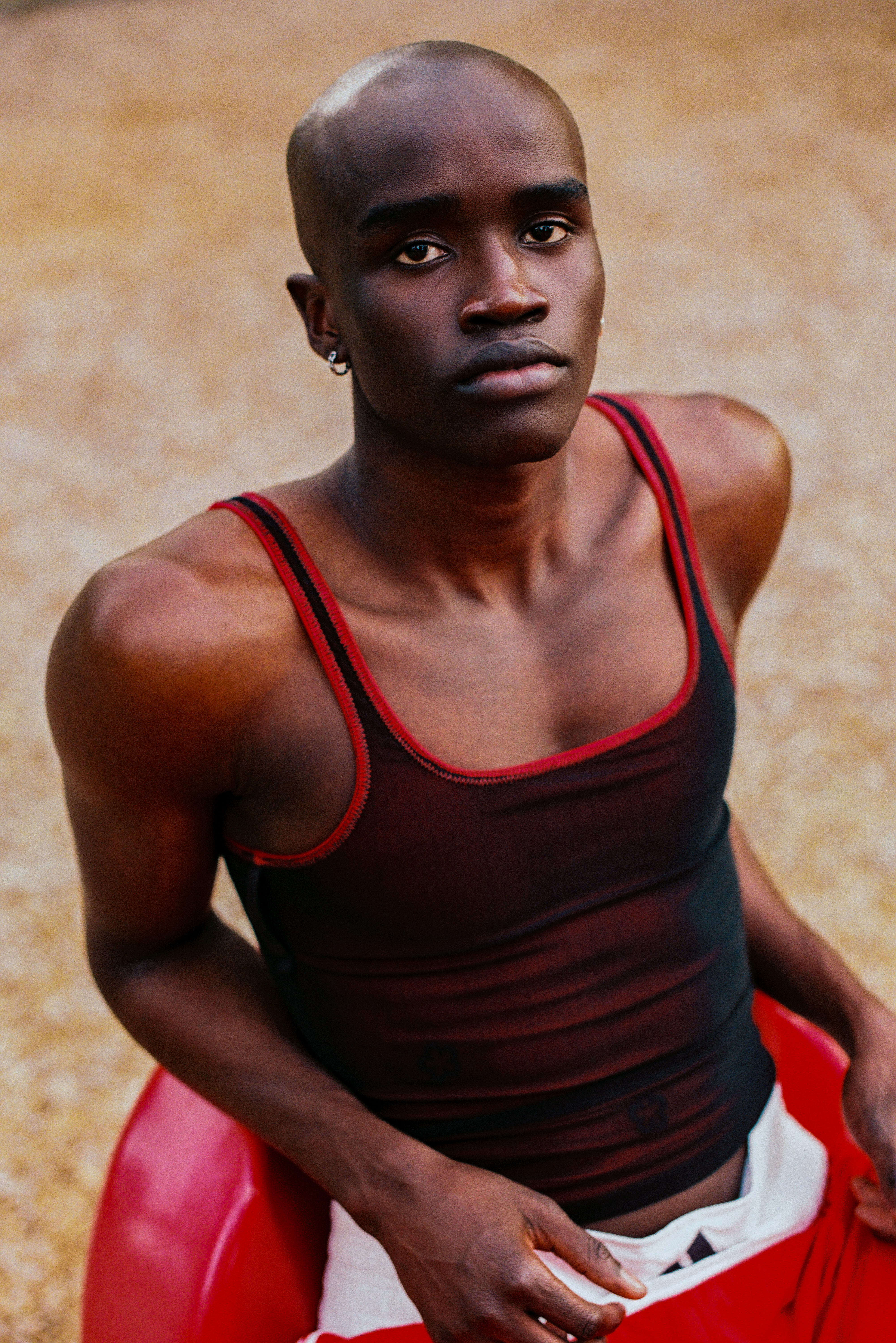 Adult male poses with a serious expression while seated in a red object outdoors. He wears a black tank top and white shorts.