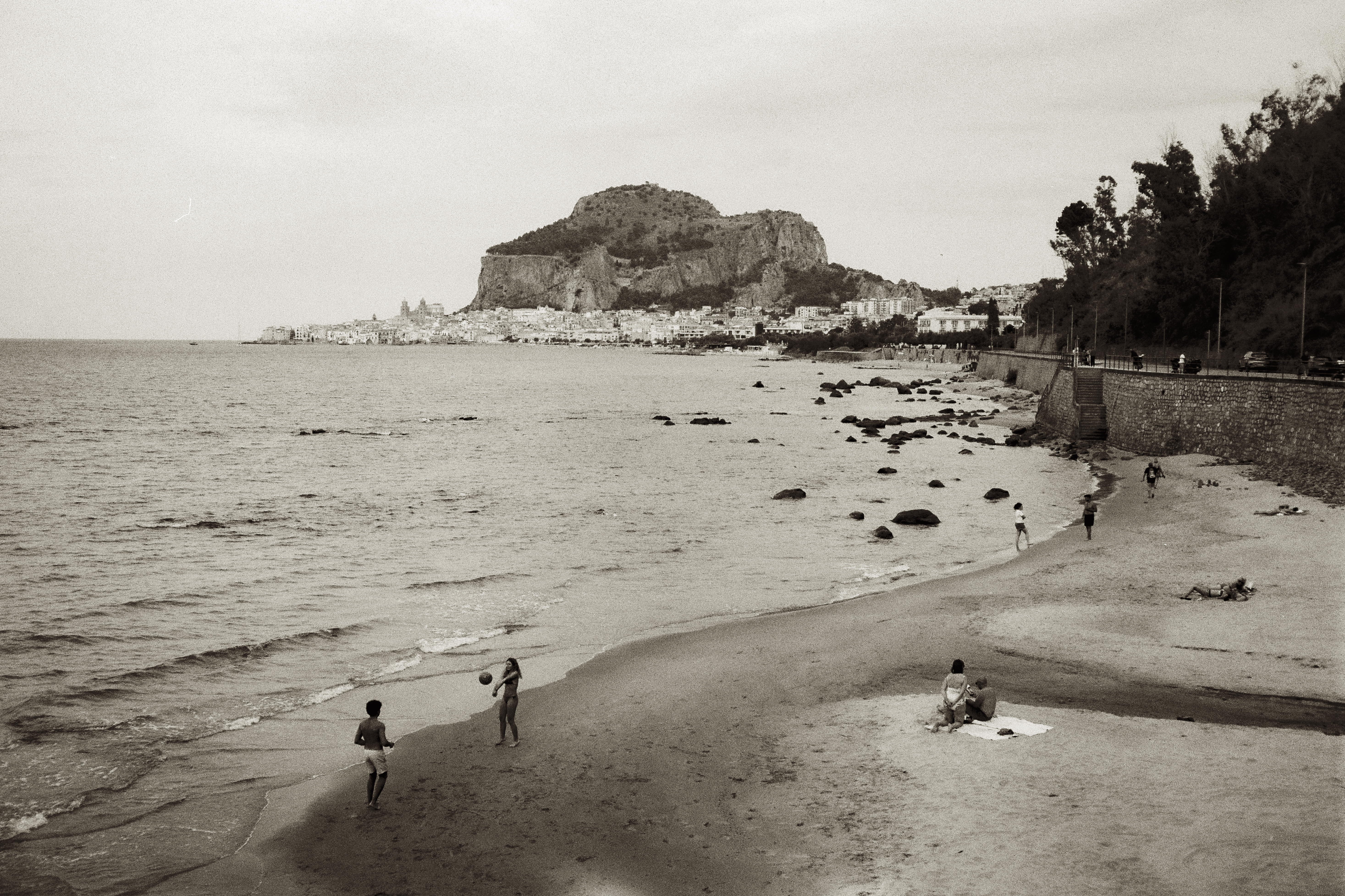 Monochrome beach scene features adults and children on sand near ocean, with a rocky mountain backdrop and cloudy sky.