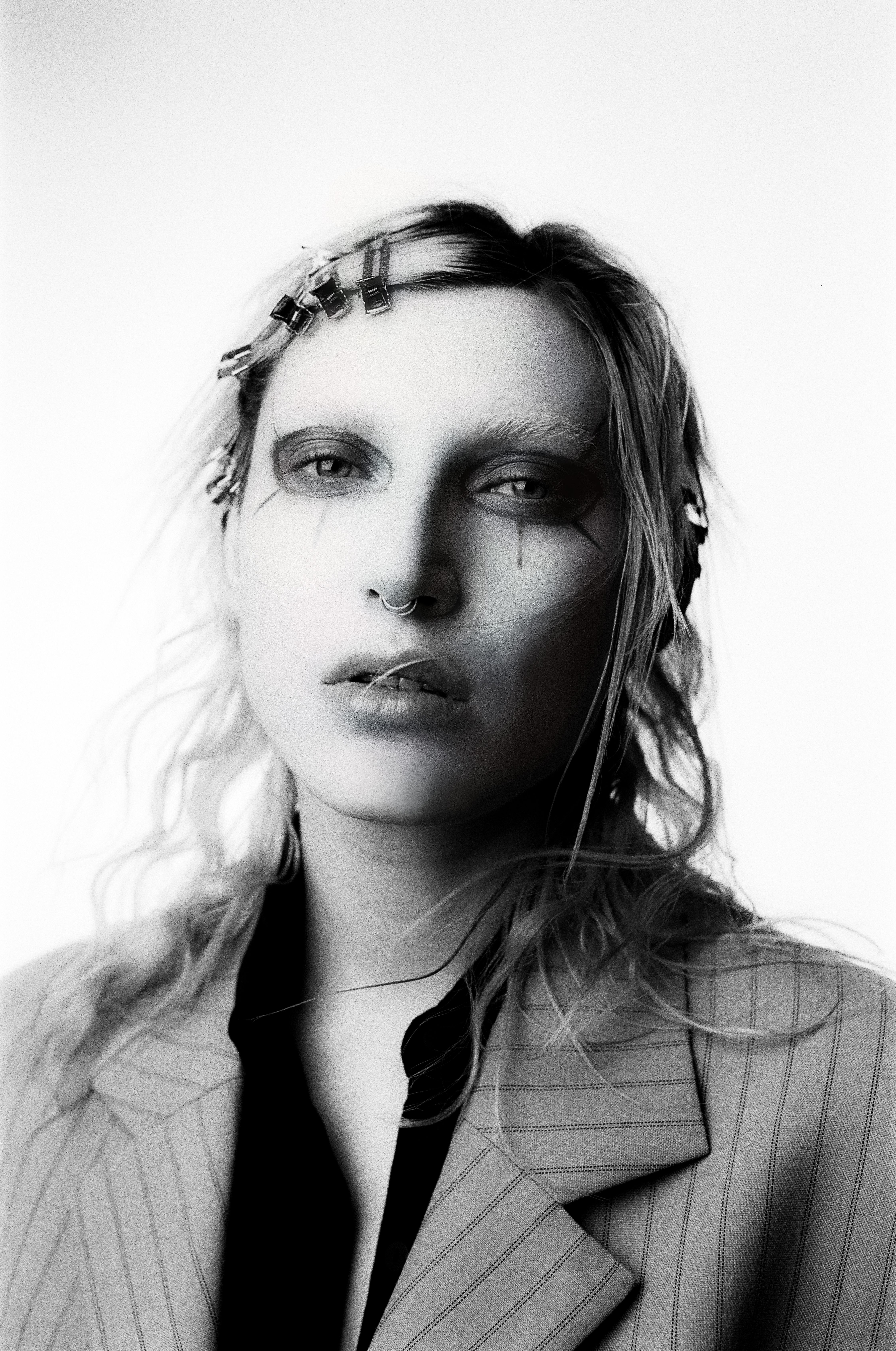 Striking black-and-white portrait of a person with creative makeup and hair clips, wearing a pinstripe blazer against a white backdrop.