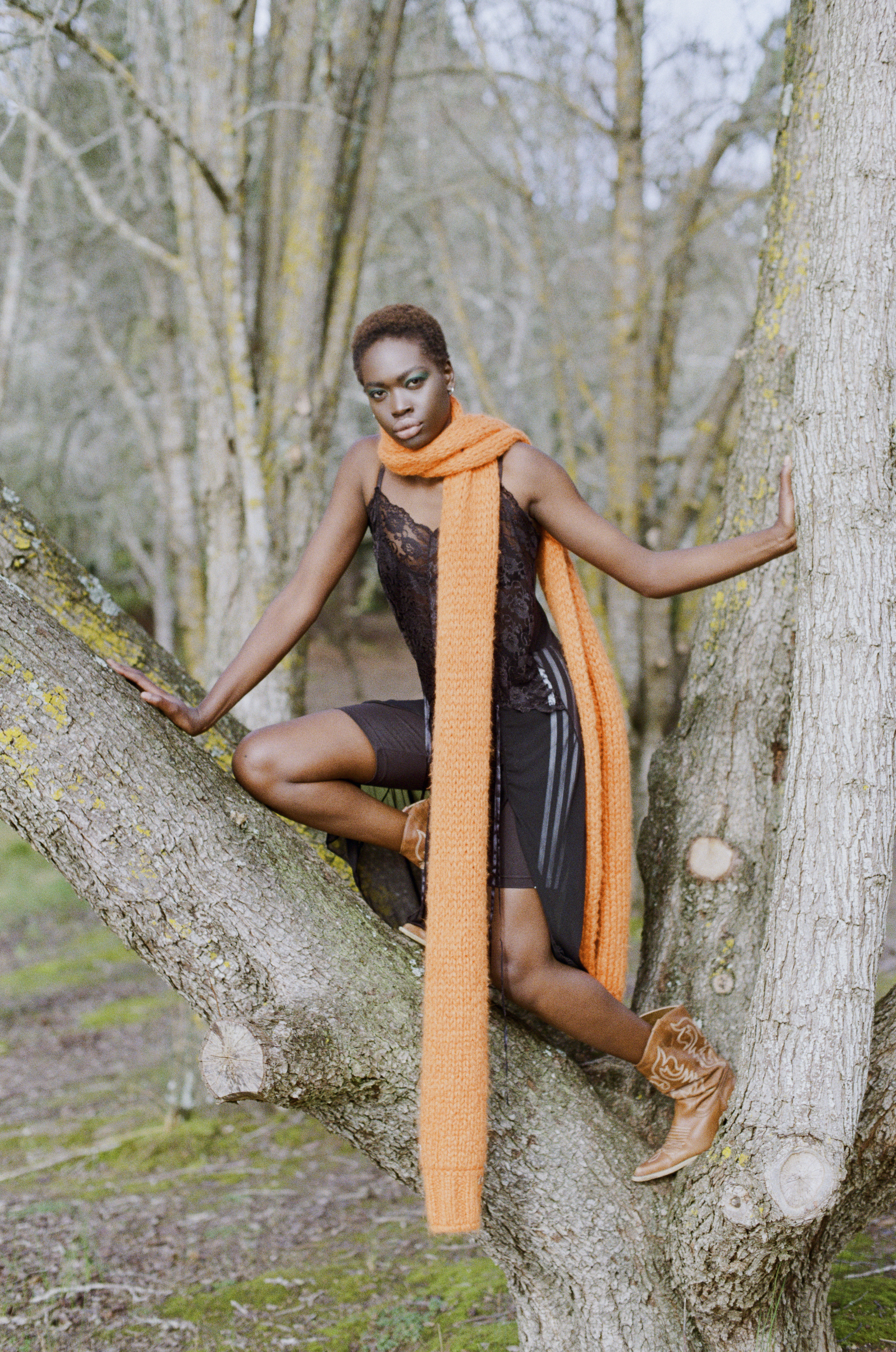 A Black woman with short hair poses in a tree wearing a black lace top, biker shorts, tan cowboy boots, and a long orange scarf.