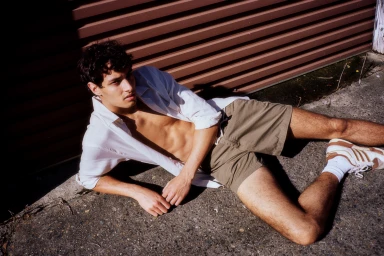 Young man lying on concrete wearing an unbuttoned white shirt and khaki shorts against a corrugated metal wall