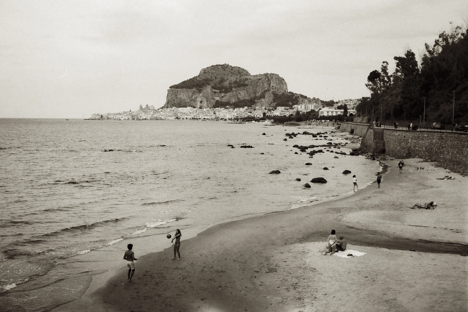 Monochrome archival photograph of a sandy beach with distant coastal town, rocky promontory, and small figures playing volleyball.