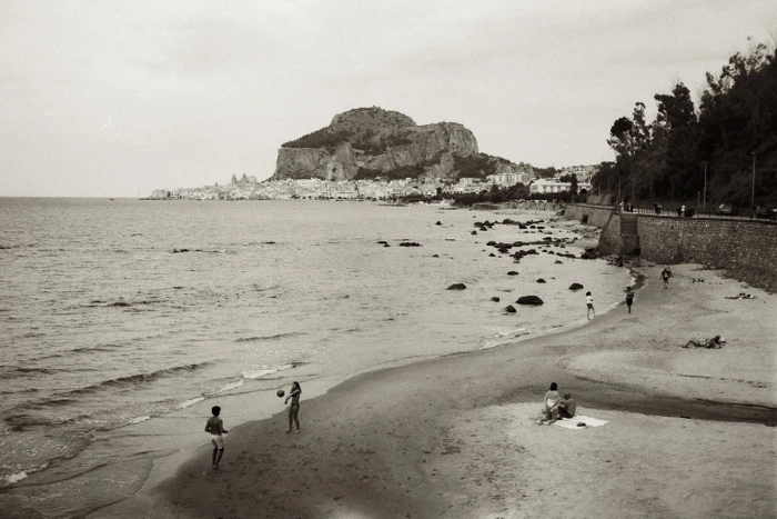 Monochrome archival photograph of a sandy beach with distant coastal town, rocky promontory, and small figures playing volleyball.