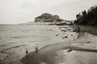 Monochrome archival photograph of a sandy beach with distant coastal town, rocky promontory, and small figures playing volleyball.