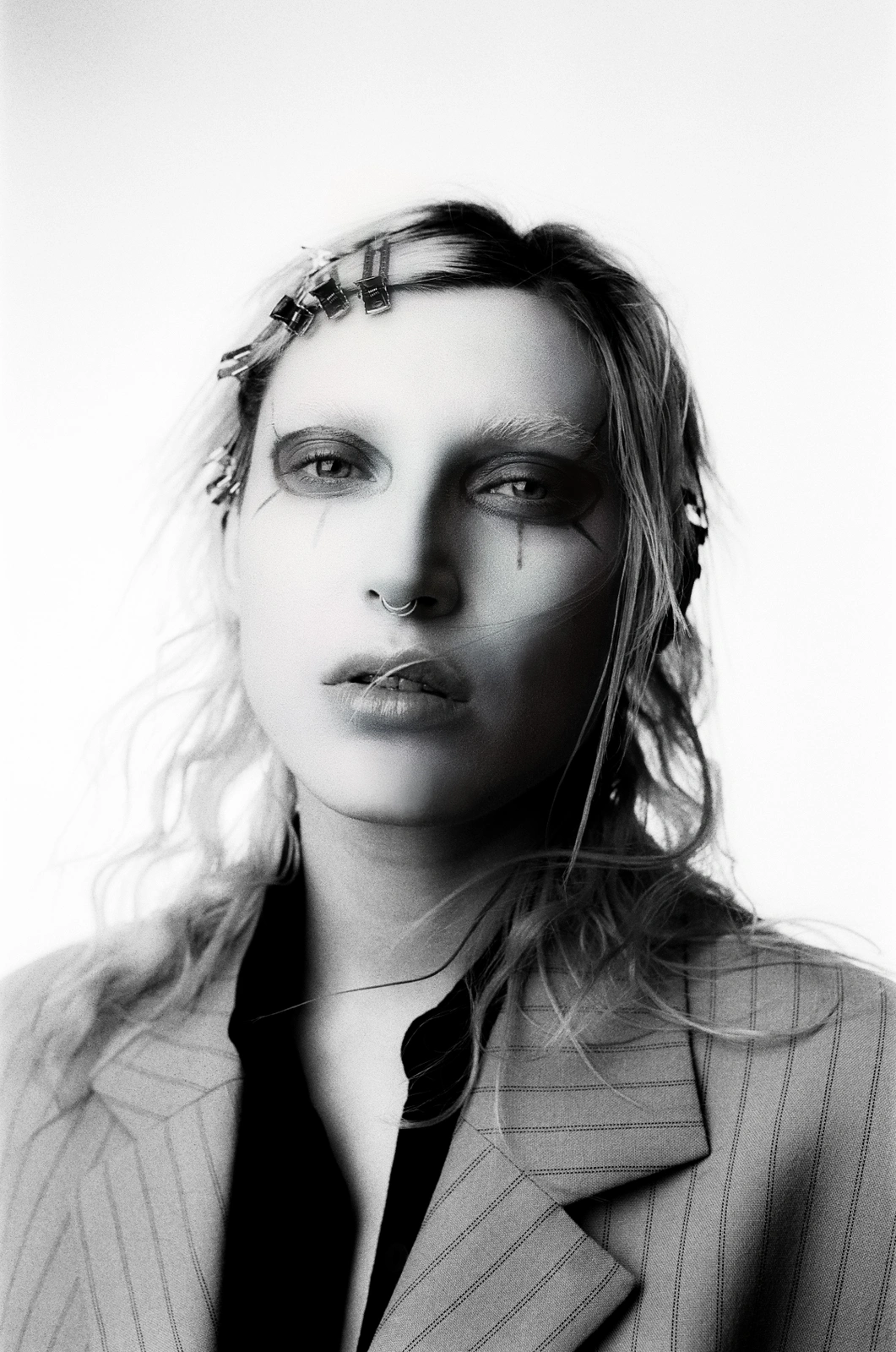Portrait of a person with dark eye makeup, nose ring, hair clips, and a striped blazer against a white background