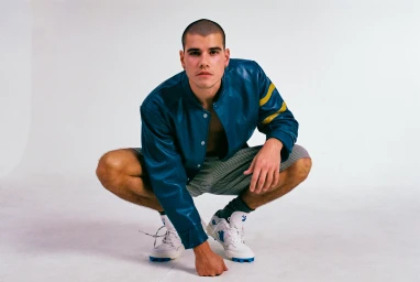 A man with a shaved head wearing a blue leather jacket and sneakers crouching against a white background.