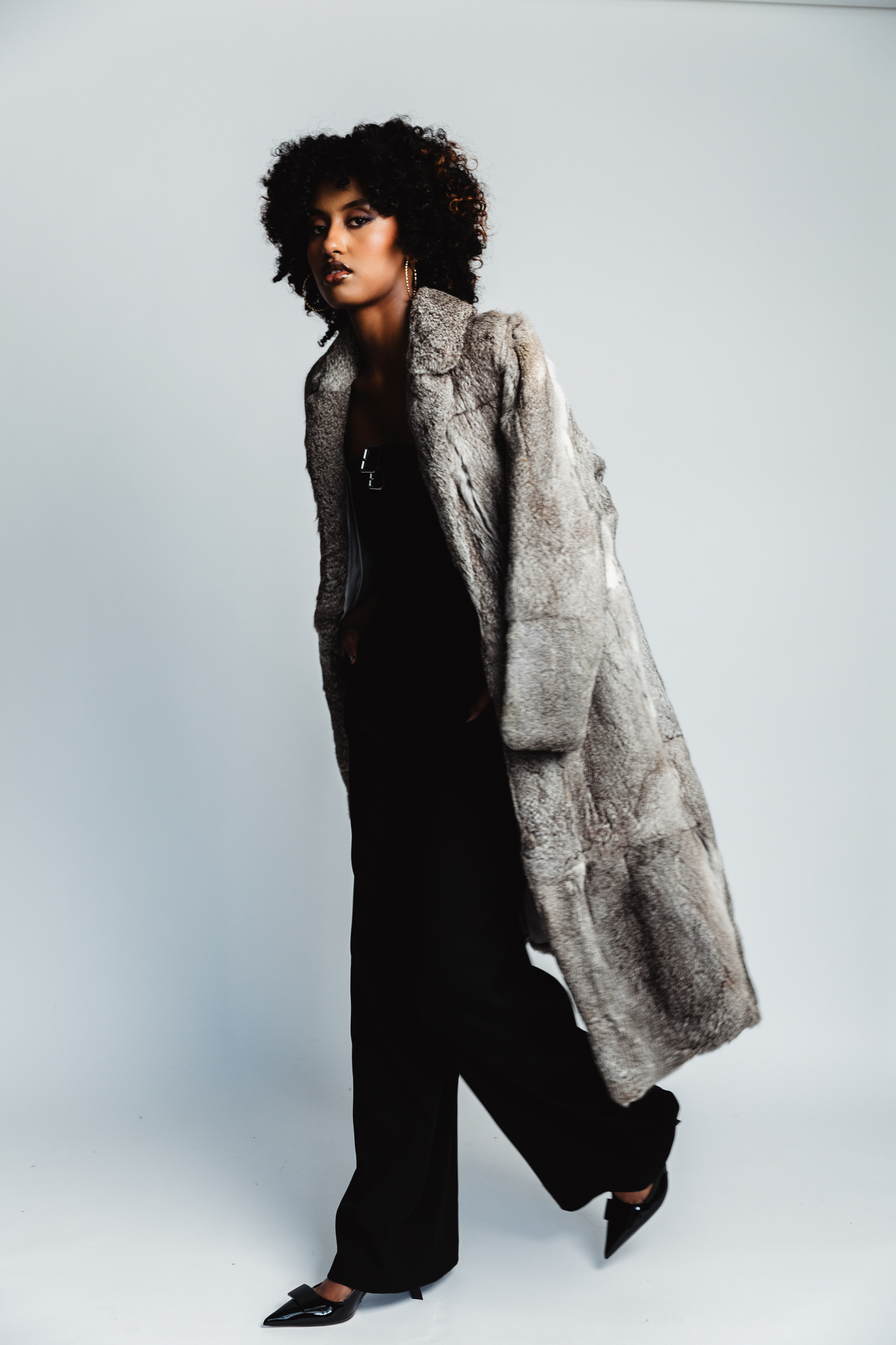 Stylish woman in a full-length fur coat, black pants, and pointed-toe shoes, posing against a soft grey background in a studio setting.