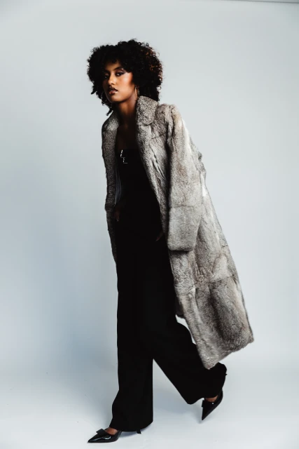 Stylish woman in a full-length fur coat, black pants, and pointed-toe shoes, posing against a soft grey background in a studio setting.