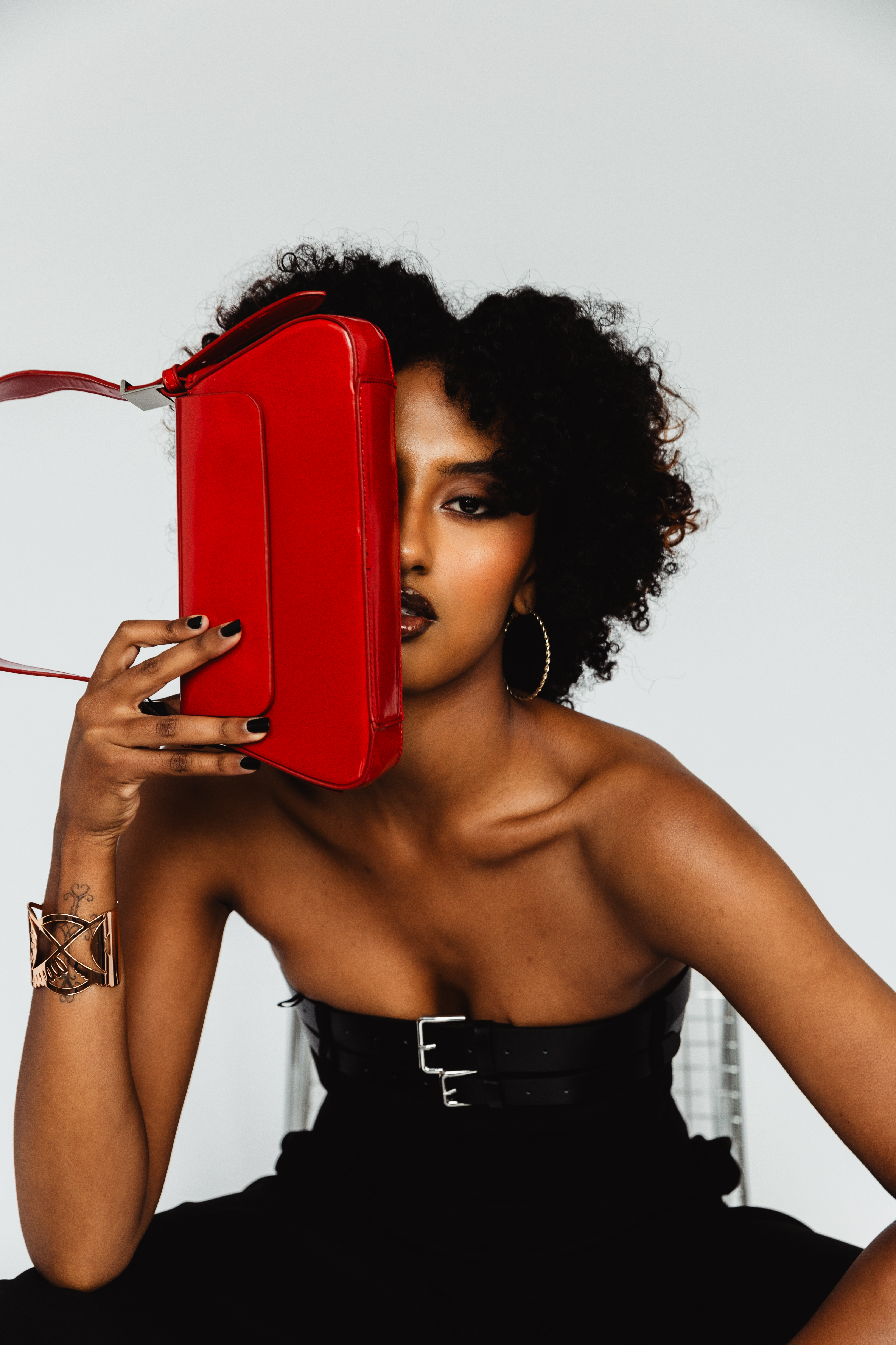 Portrait of a woman posing with a red patent leather handbag covering half of her face, wearing a black strapless dress and hoop earrings.