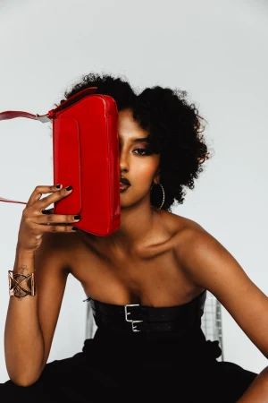 Black woman holding a glossy red handbag up to her face against a white seamless studio background