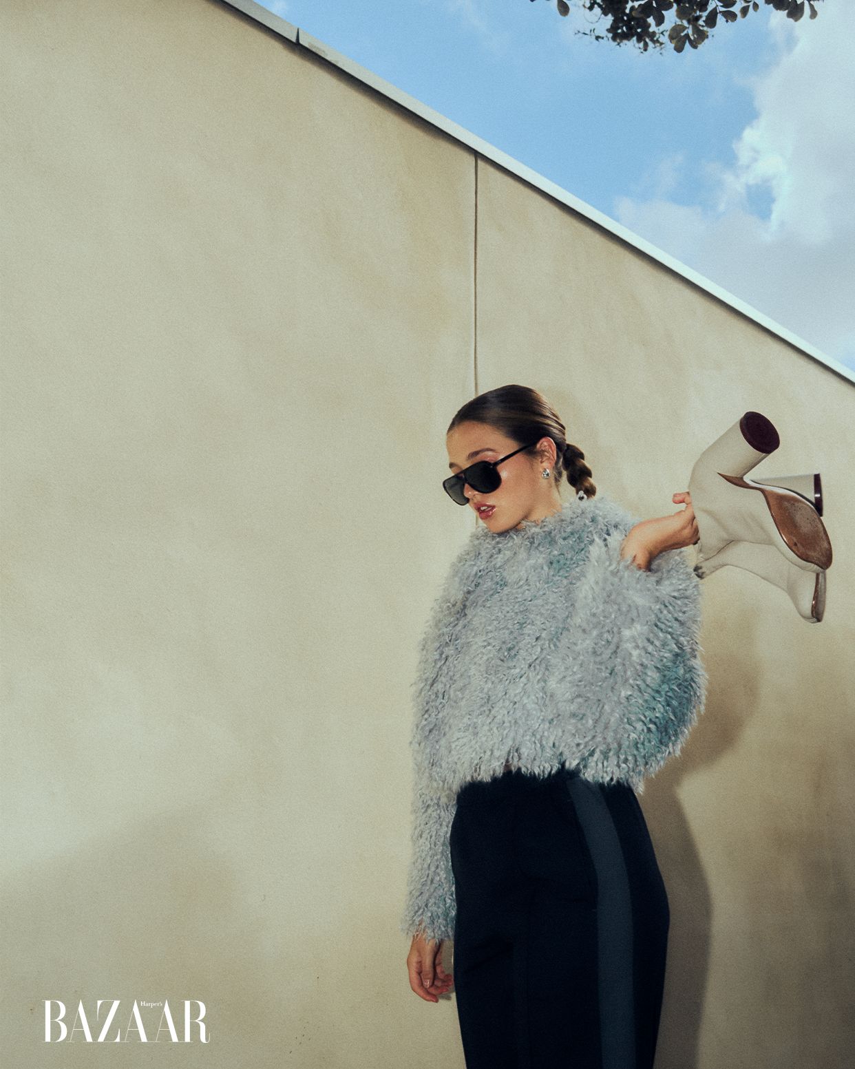 Fashion shot of a woman wearing a grey furry jacket, black skirt and sunglasses, holding beige boots, posing against a neutral wall and sky.