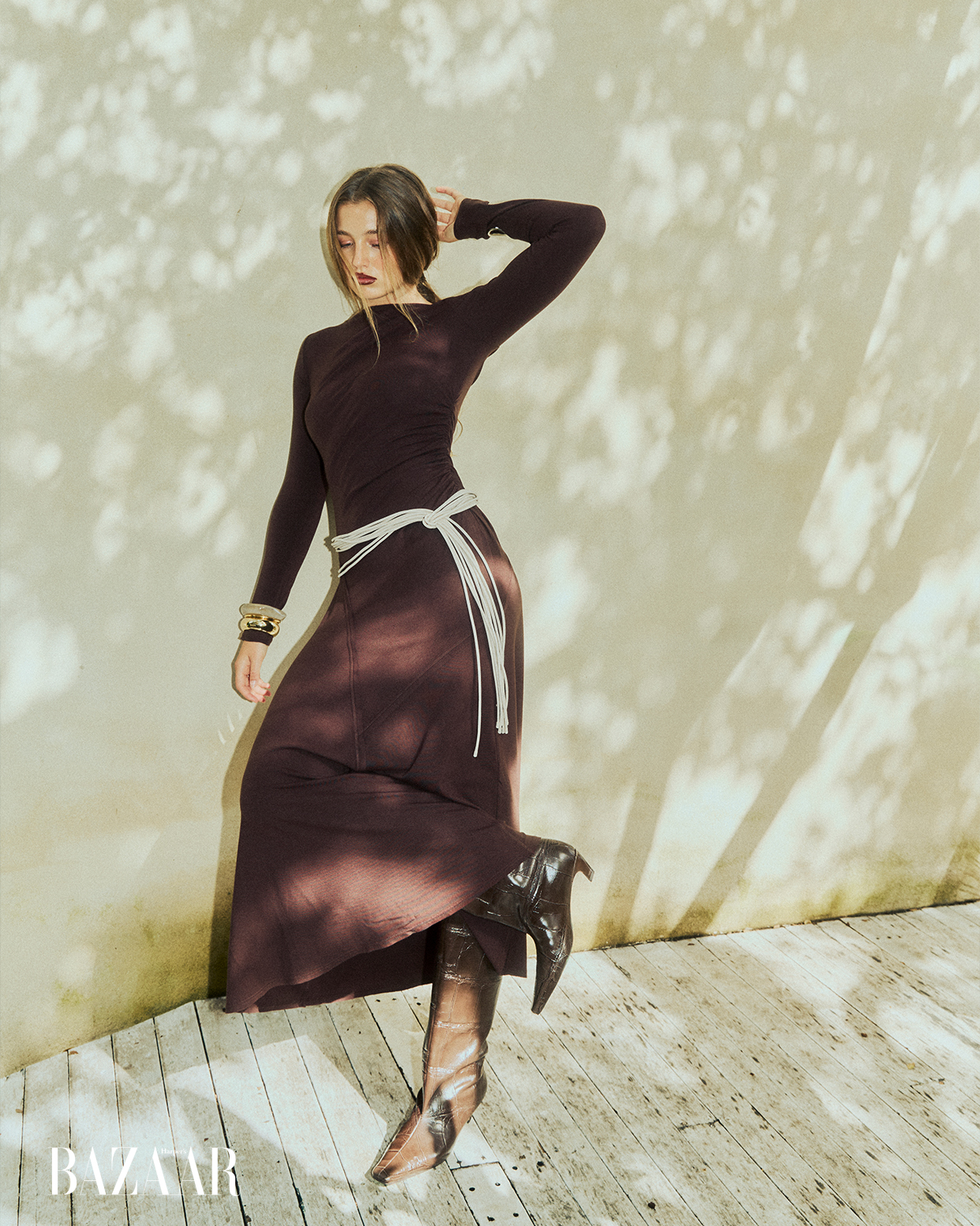 Elegant adult female poses in long-sleeve brown dress with tan fringe belt and black cowboy boots against a light wall with leaf shadows.