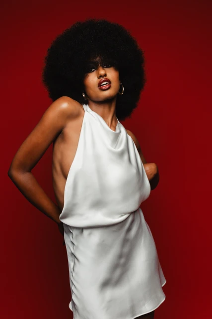 Glamorous woman with an afro poses confidently against a red backdrop in a fashionable white mini dress and gold hoop earrings.