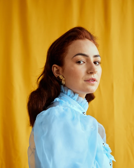 Portrait of a beautiful woman with auburn hair, wearing a light blue ruffled blouse and gold earrings, posing against a yellow backdrop.