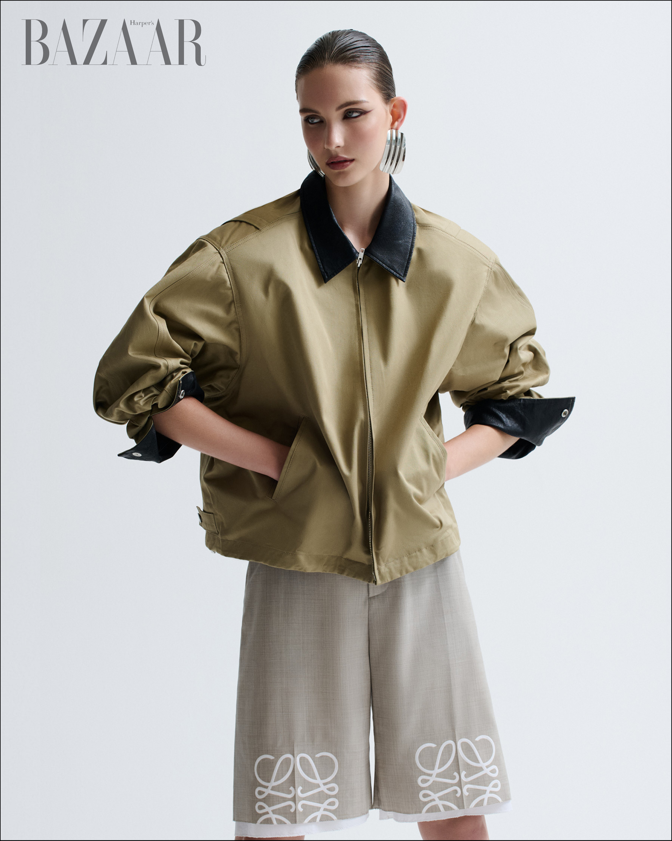 Fashion editorial shot of a young woman wearing a tan jacket with black collar and grey shorts, posing against a neutral background.