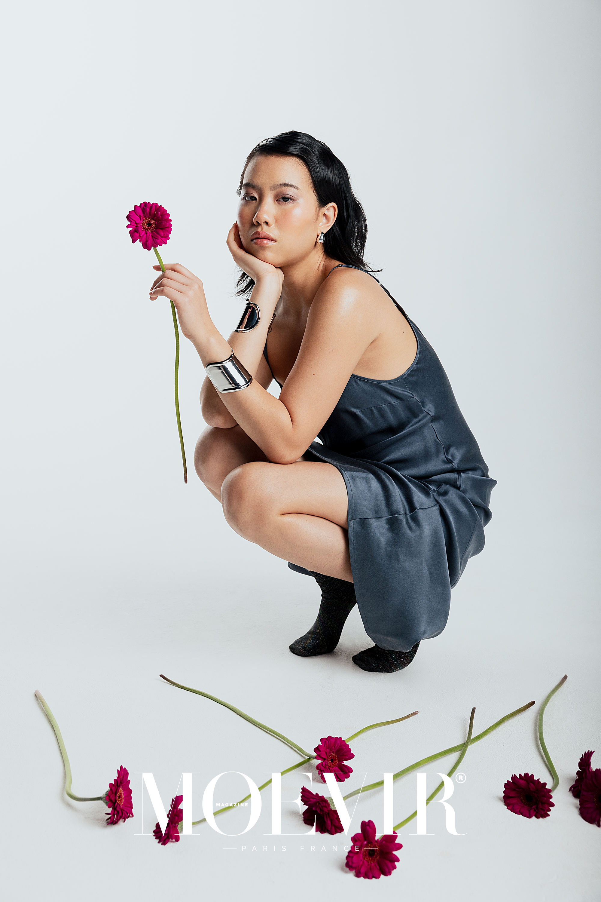 An Asian woman poses in a studio wearing a blue dress and silver cuffs, holding a pink flower, surrounded by scattered flower heads.