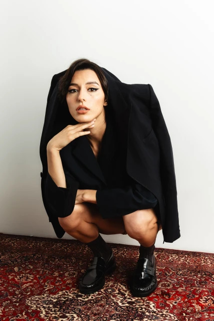 Woman in black blazer poses crouching indoors on ornate red rug. She wears winged eyeliner and patent leather loafers against white wall.