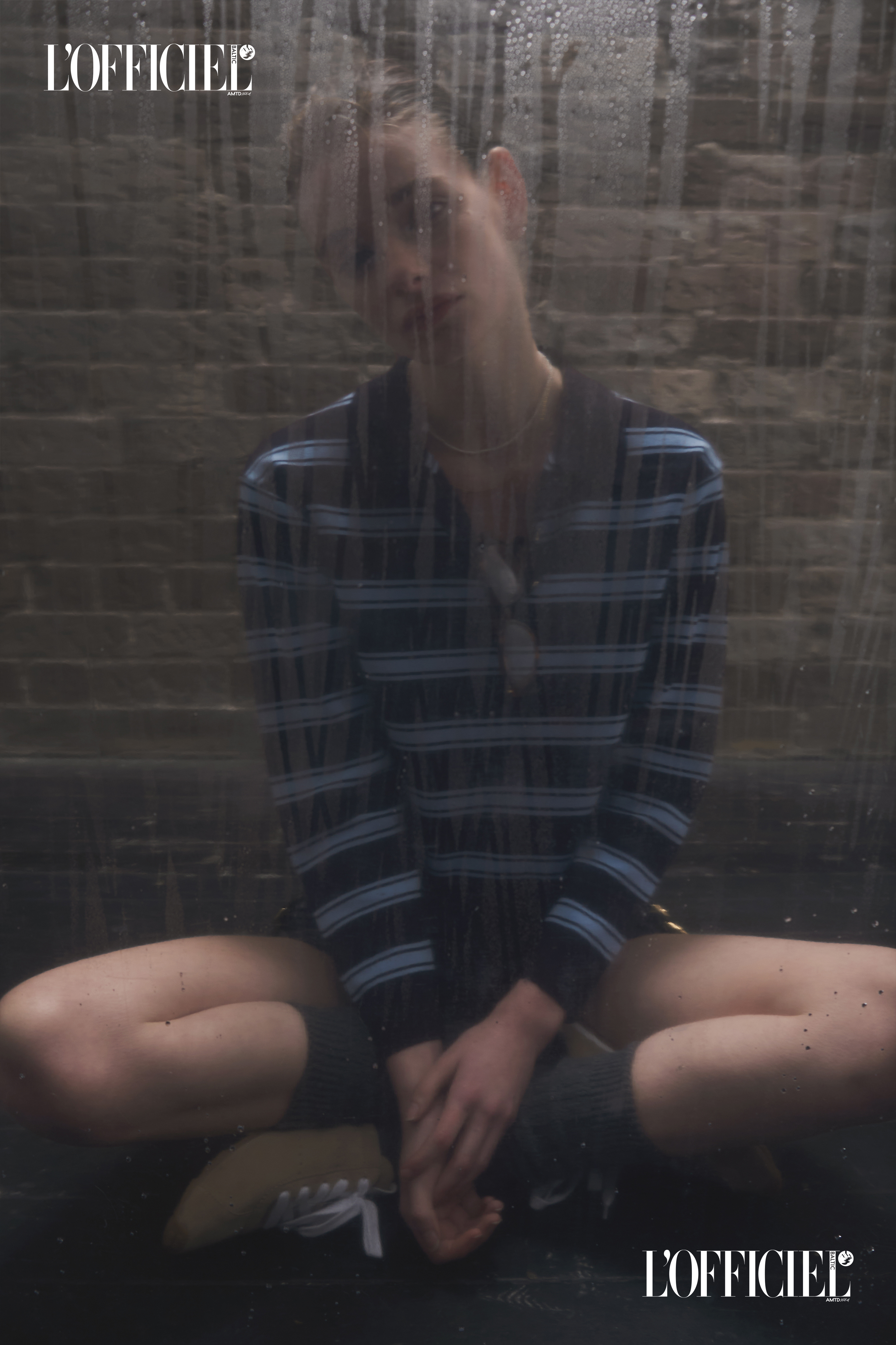 Fashion editorial shot of a young adult female model in a striped shirt and shorts, sitting against a neutral brick background with water droplets.